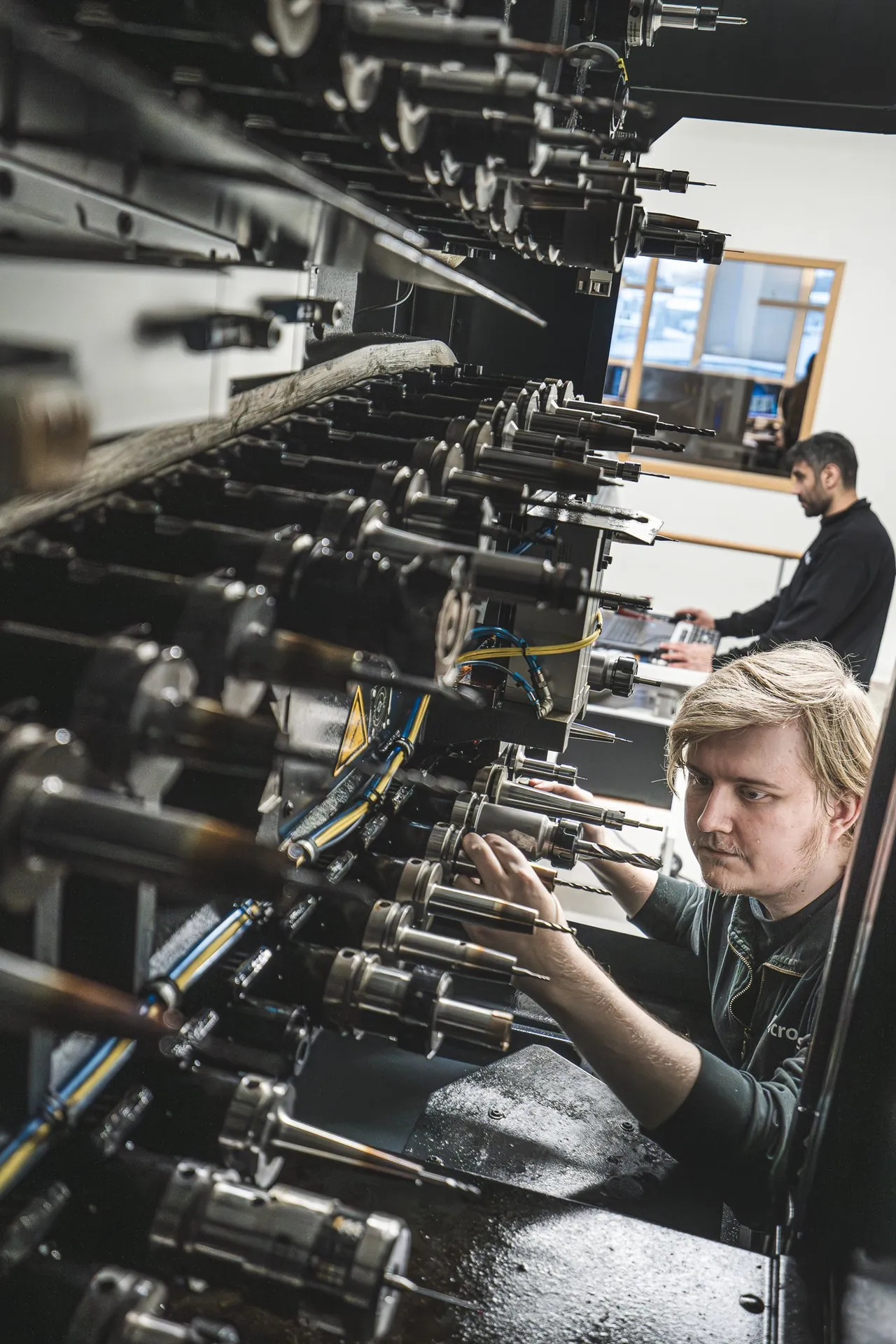 Operator loading tools into a large tool magazine while a colleague works at a computer in the background