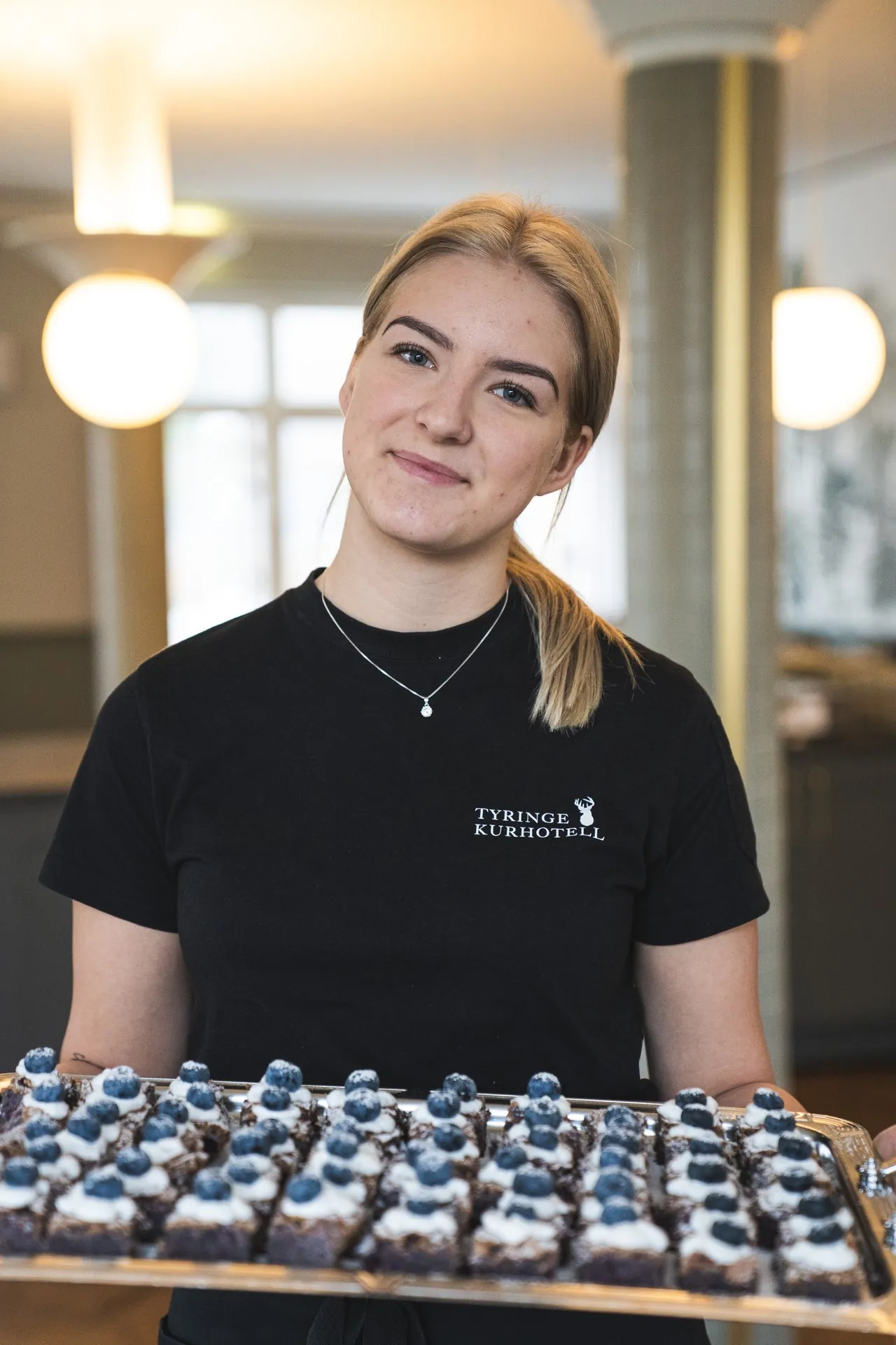 Servitris med Tyringe Kurhotell-tröja bär bricka med blåbärsbakelser i lobbyn
