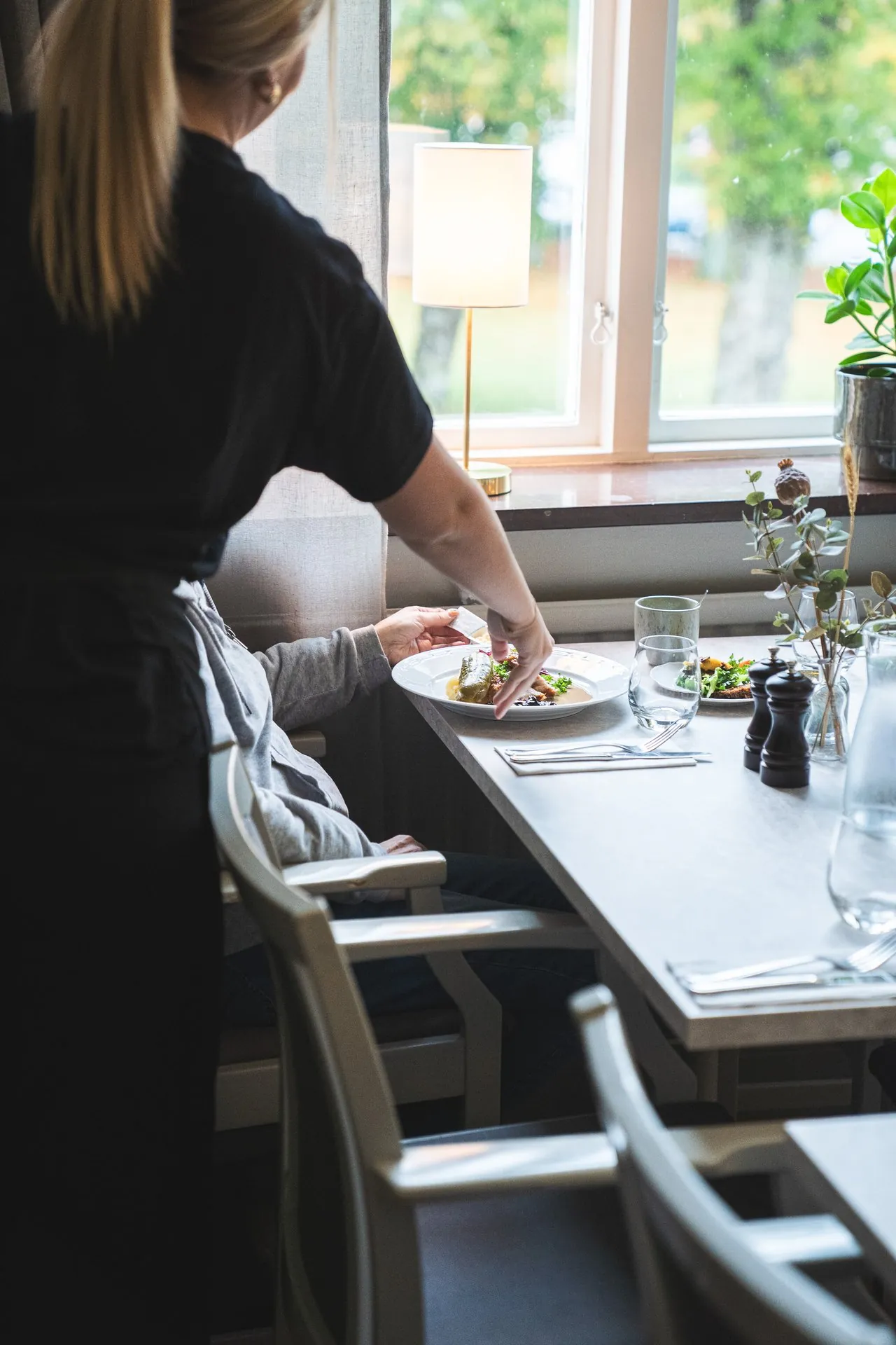 Servitris serverar lunch vid fönsterbord med grönska utanför