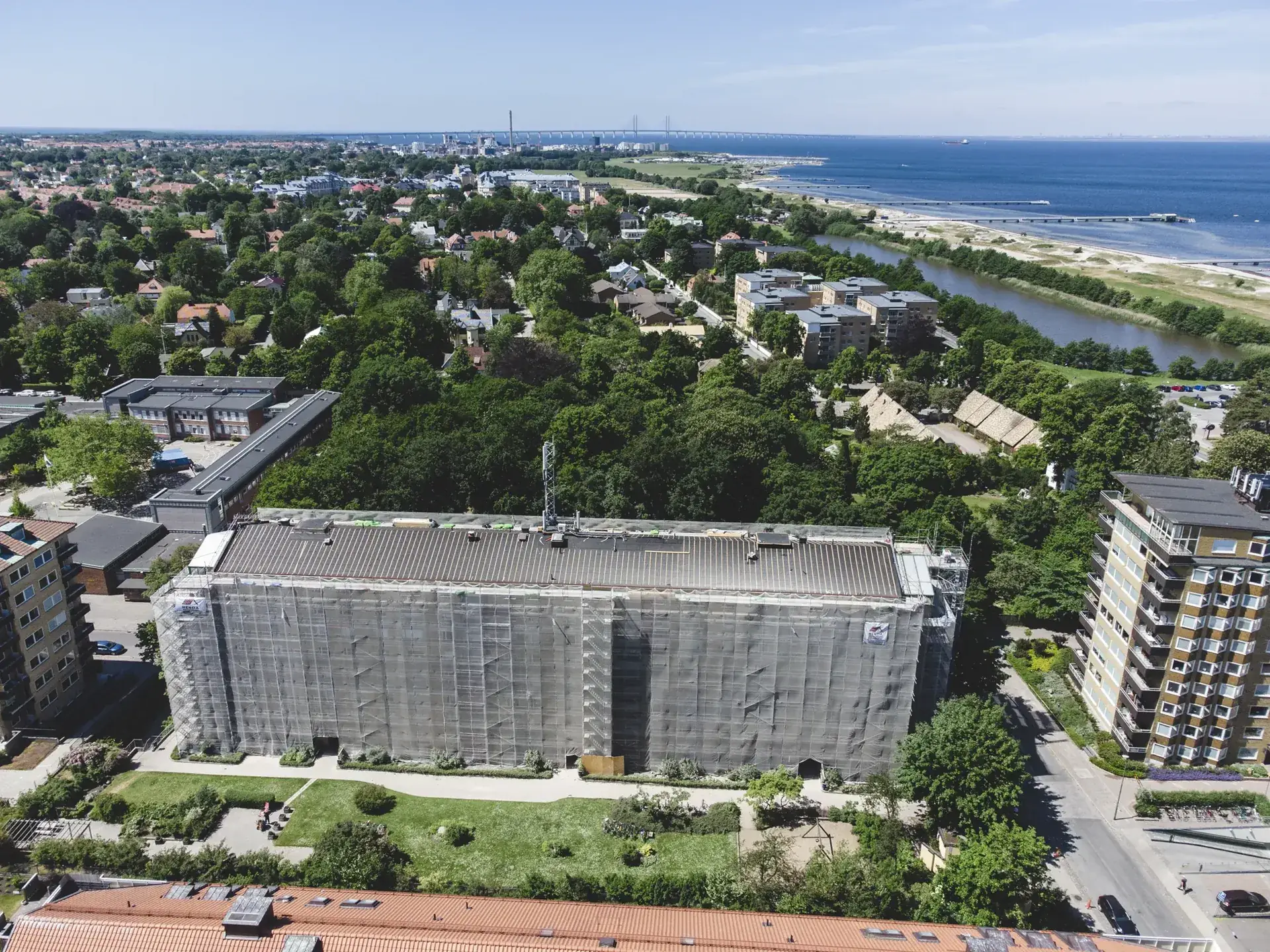 Flygfoto över ett inplastat flerbostadshus under fasadrenovering i Malmö, med kusten och Öresundsbron i bakgrunden