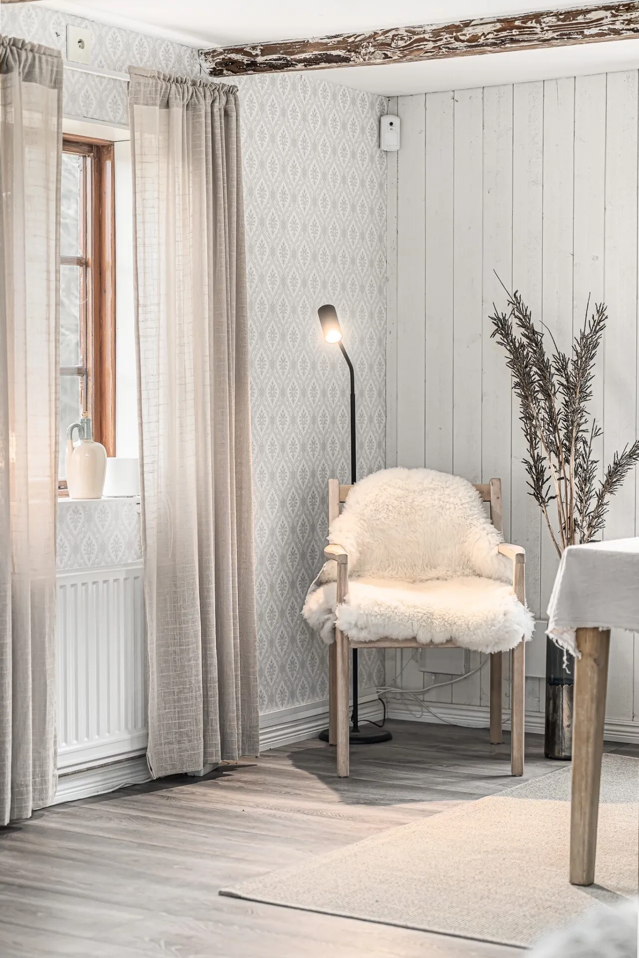 Stillsam vrå med fårskinnsklädd stol och linnegardin i äldre hus åt HusmanHagberg